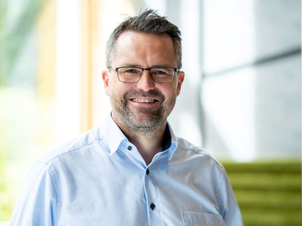 Ein Mann mit Brille und hellblauem Hemd lächelt in einem Innenraum in der Nähe großer Fenster.