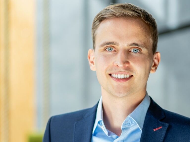 Ein Mann in einer blauen Anzugsjacke lächelt, während er in einem Haus in der Nähe eines Fensters steht.