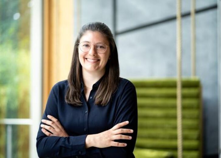 Eine Frau mit Brille und langen Haaren steht mit verschränkten Armen in einem Haus und lächelt.