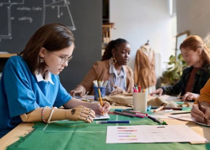 Vier Personen arbeiten gemeinsam an einem Tisch in einem Klassenzimmer an Stoffen und Skizzen.