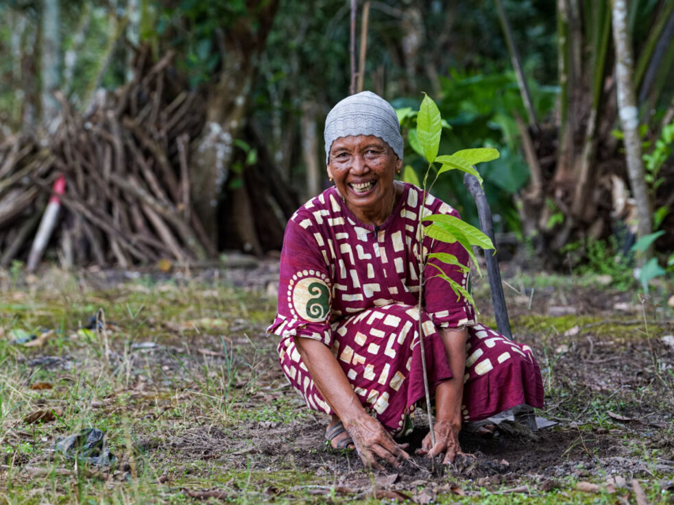 Eine lächelnde Frau in einem gemusterten Kleid pflanzt einen kleinen Bäumchen im Freien.
