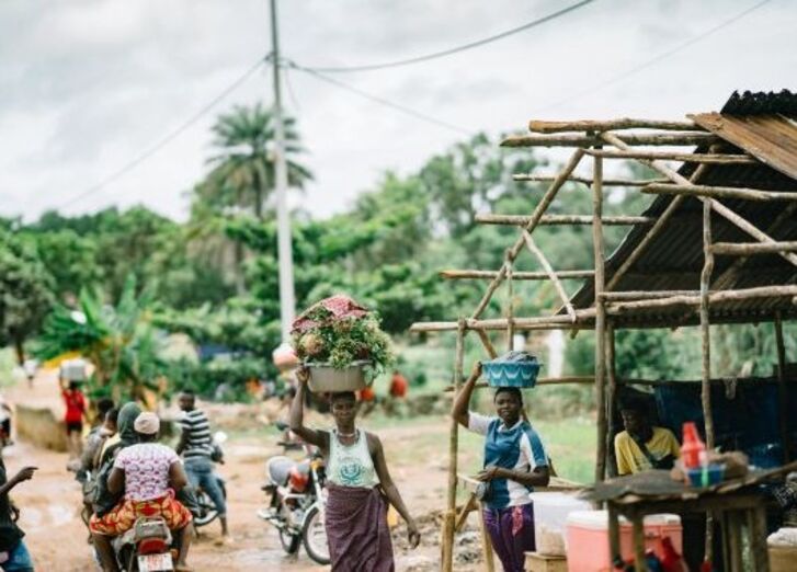 Frauen tragen Waren auf ihren Köpfen in der Nähe einer Holzkonstruktion in einem ländlichen Gebiet.