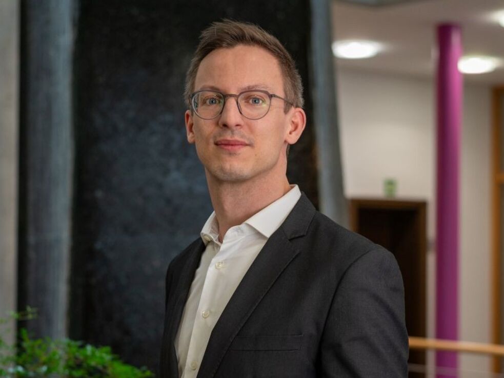 Ein Mann im Anzug steht in einem Haus in der Nähe einer Pflanze und schaut in die Kamera.